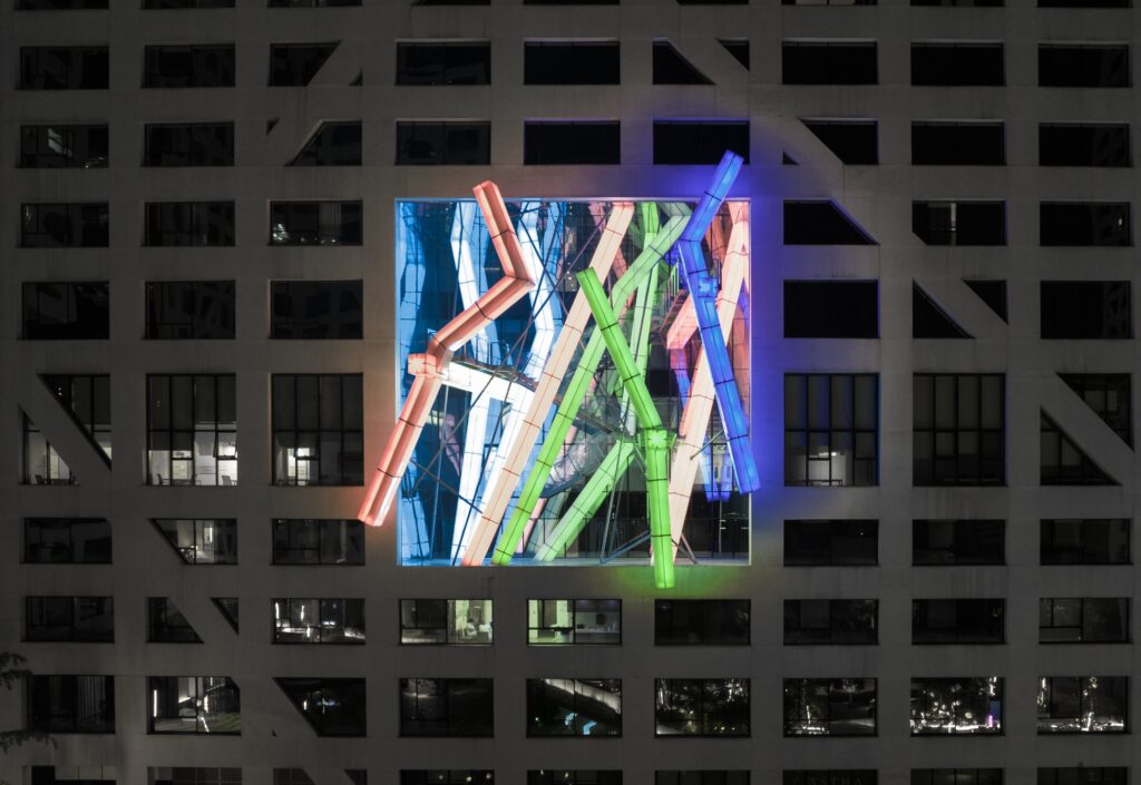 Night scene of a gray office building with a glass atrium in the center, lit by colorful neon tubes in red, blue, green and purple.