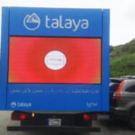 Talaya 1 Rear view of a blue Talaya advertising truck with a large red screen and a white circular logo, parked on a street.