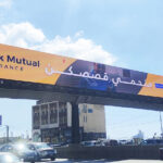 vivad jal eldib2 Large yellow-orange billboard over a highway reading 'Rock Mutual Insurance' with a blue logo, Arabic text, and a blue sky background, indicating an insurance ad.
