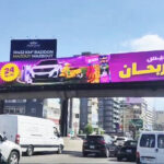 vivad jal eldib8 Bright purple billboard spanning a city highway, with cars driving beneath and Arabic text on the ad banner.