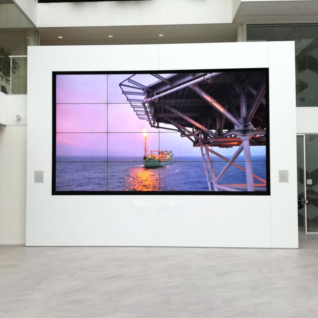 Large wall-mounted screen shows an offshore oil platform with a lit flare and a support ship on the sea at sunset.