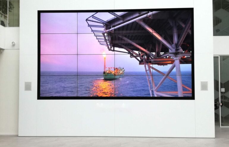 Large wall-mounted screen shows an offshore oil platform with a lit flare and a support ship on the sea at sunset.